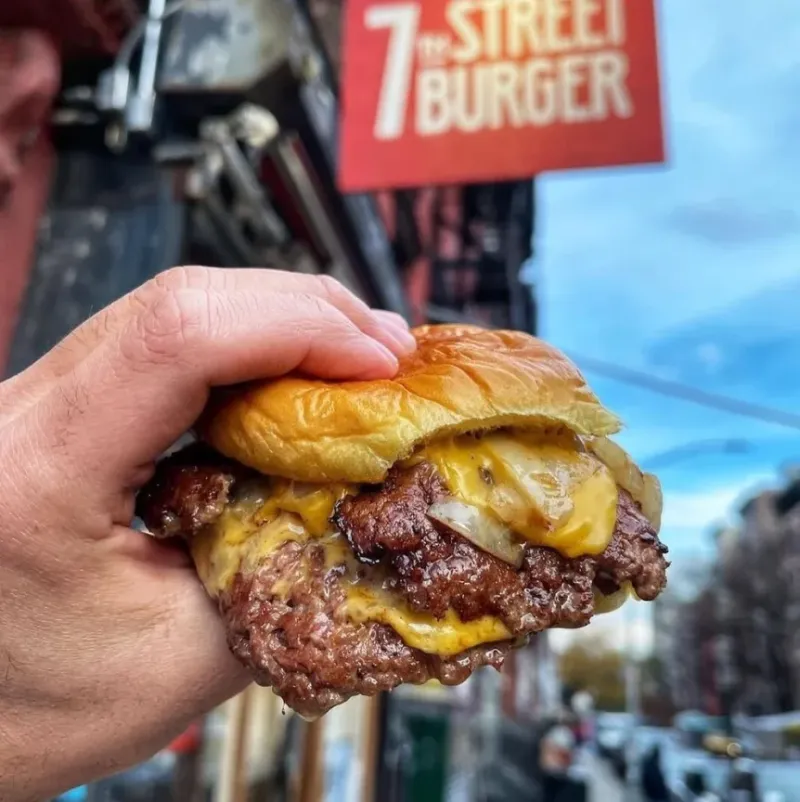 Hand holding a 7th Street Burger double cheeseburger outside a restaurant storefront